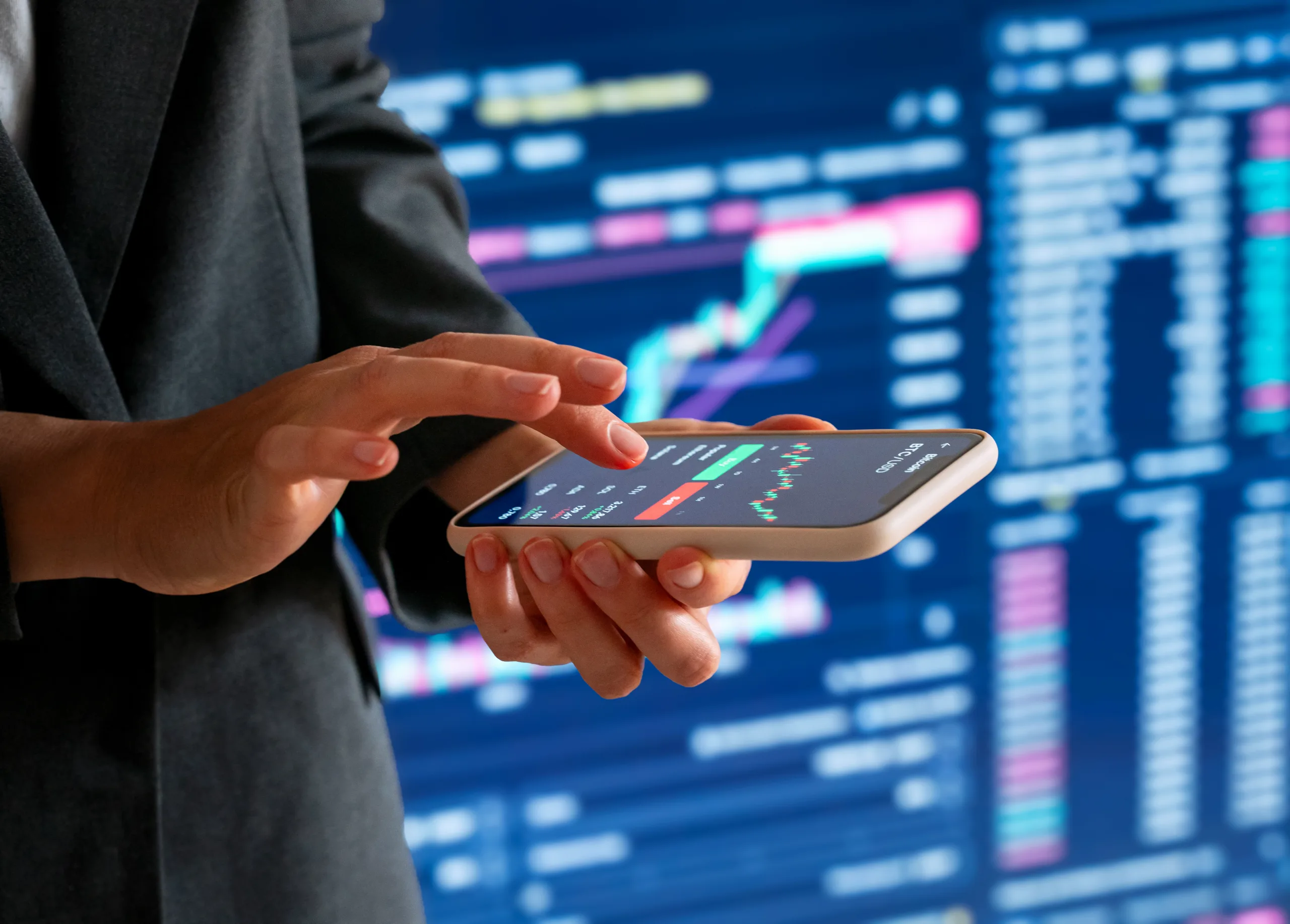 Female investor using smartphone for crypto trading in front of electronic chart
